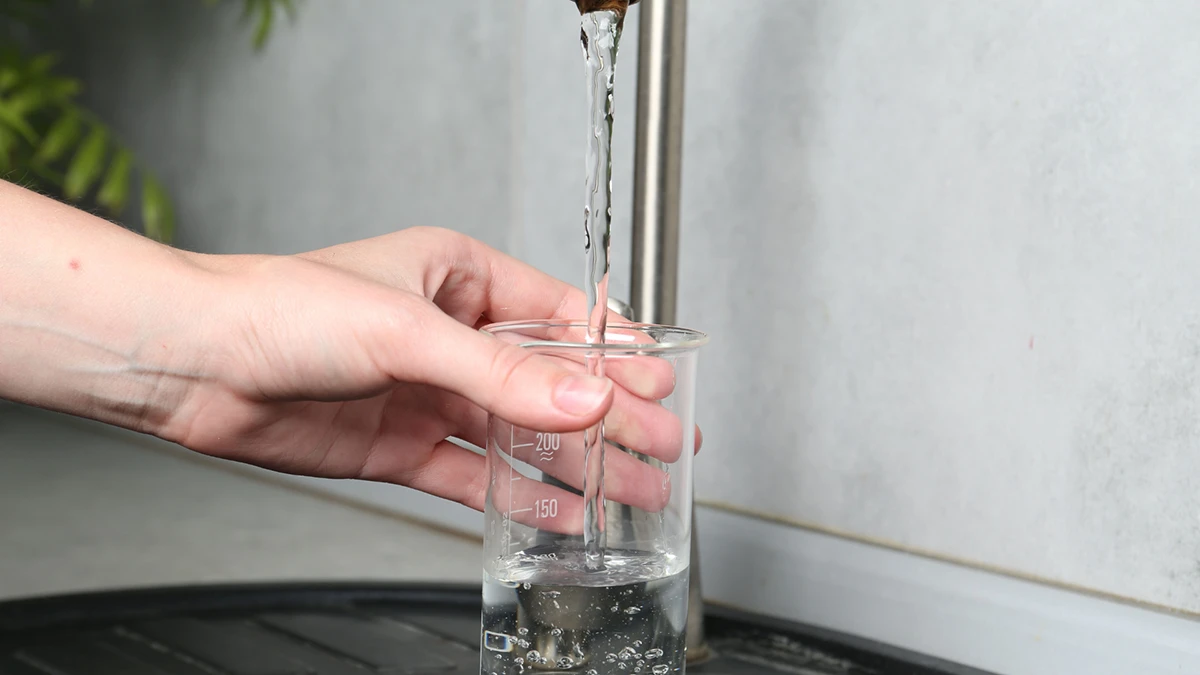 A hand holds a glass beaker under a running faucet, filling it with water. The setting is a clean, tiled kitchen or lab, creating a simple, focused mood.