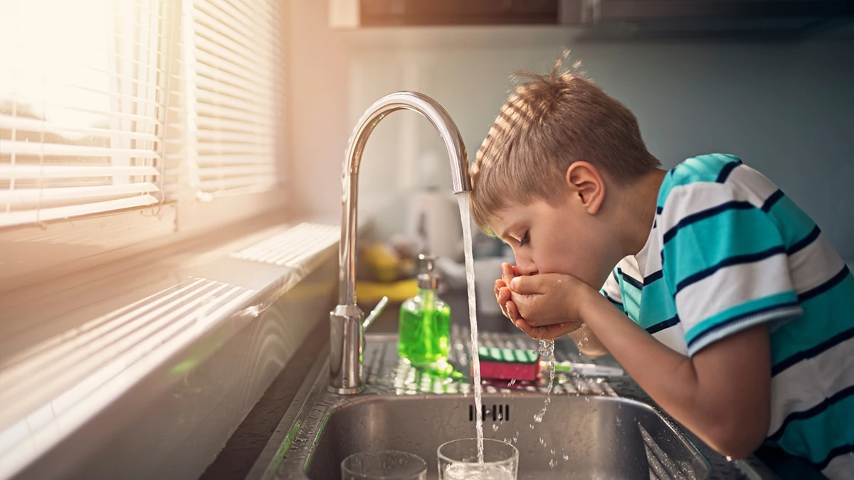 A young boy drinks water from a kitchen sink, smiling as he enjoys a refreshing sip.
