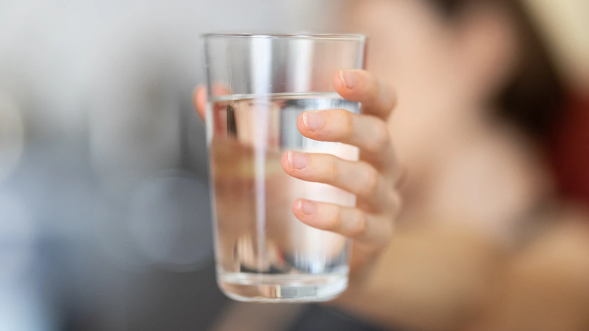 A hand holding a clear glass of water against a soft, blurred background. The image conveys a sense of refreshment and hydration.