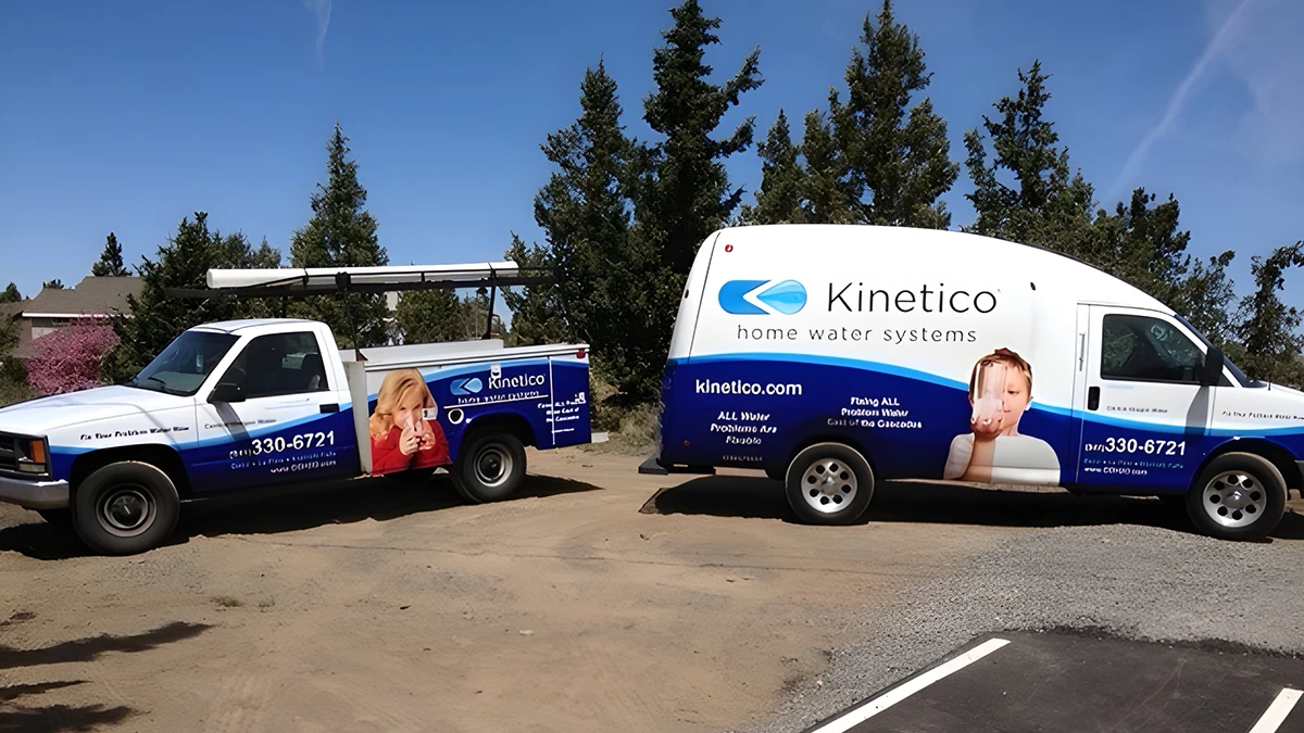 Two utility vehicles with "Kinetico Home Water Systems" logos are parked on a dirt road. The van features a child's face, and trees are in the background.