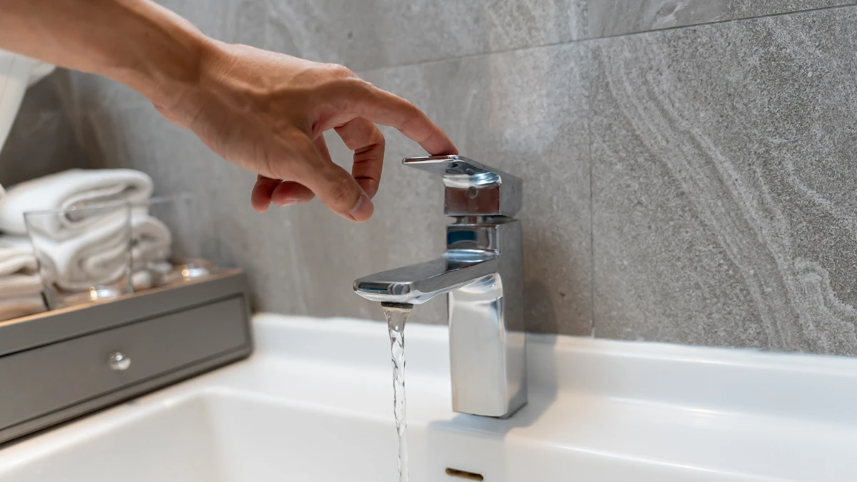 A hand reaches to turn on a modern chrome faucet, with water flowing into a white bathroom sink. Gray stone backsplash and folded towels are visible.