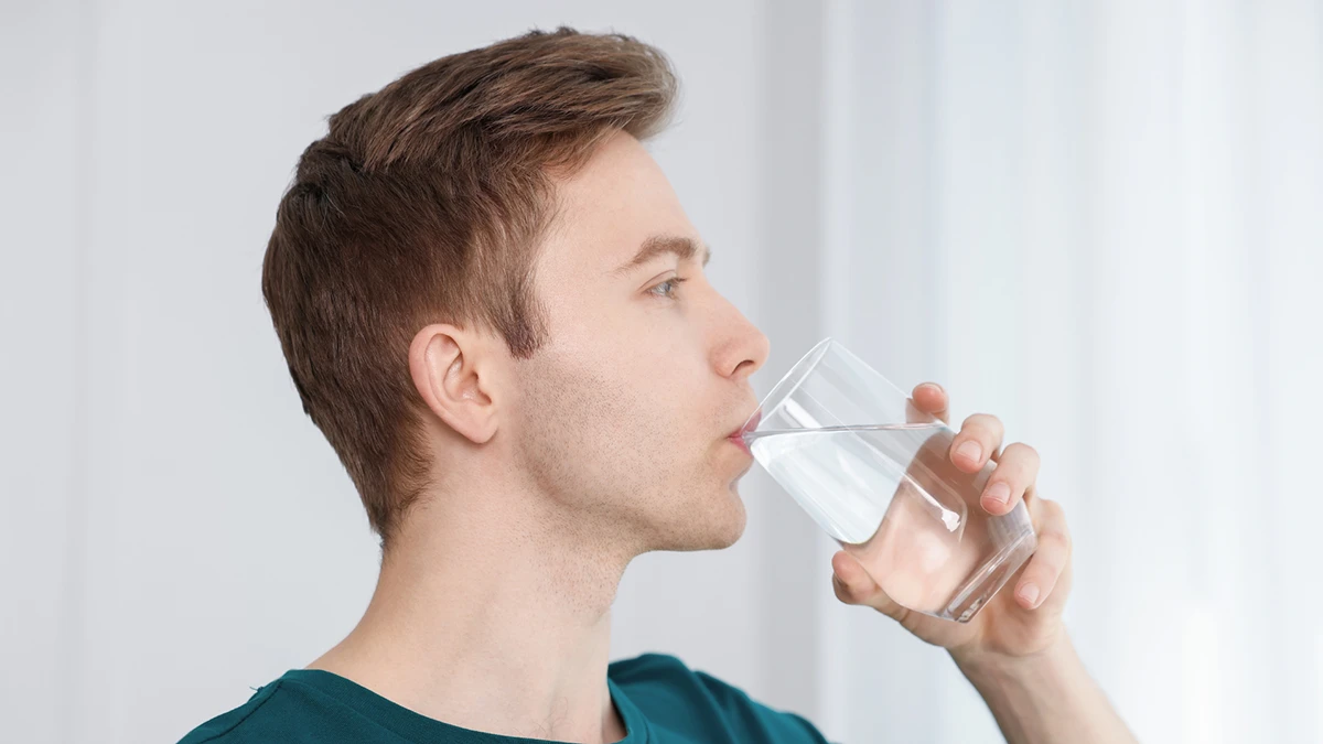 A man with short brown hair is drinking water from a clear glass, set against a softly lit interior background. He appears calm and focused. A man with short brown hair is drinking water from a clear glass, set against a softly lit interior background. He appears calm and focused.